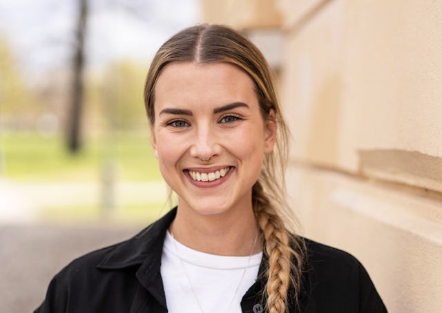En kvinna med långt blont hår i en sidofläta står utomhus nära en ljus vägg och ler.