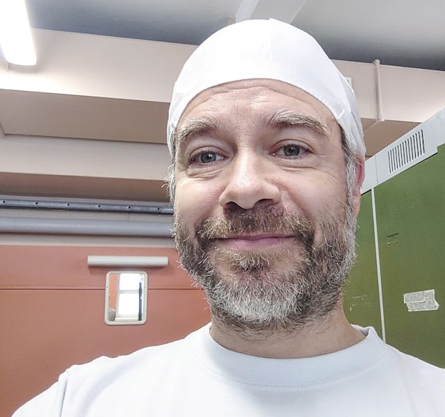 En man med skägg, vit keps och vit tröja ler mot kameran inomhus.