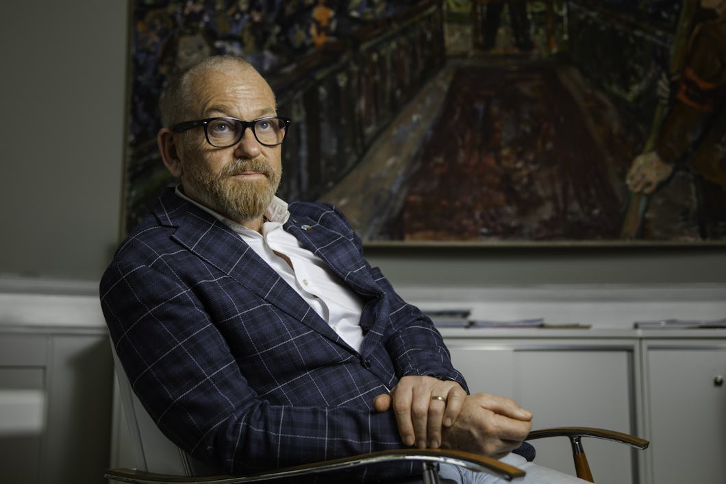 Johan Lindholm med glasögon, skägg och rutig kavaj sitter på en stol med armarna i kors framför en tavla på väggen.
