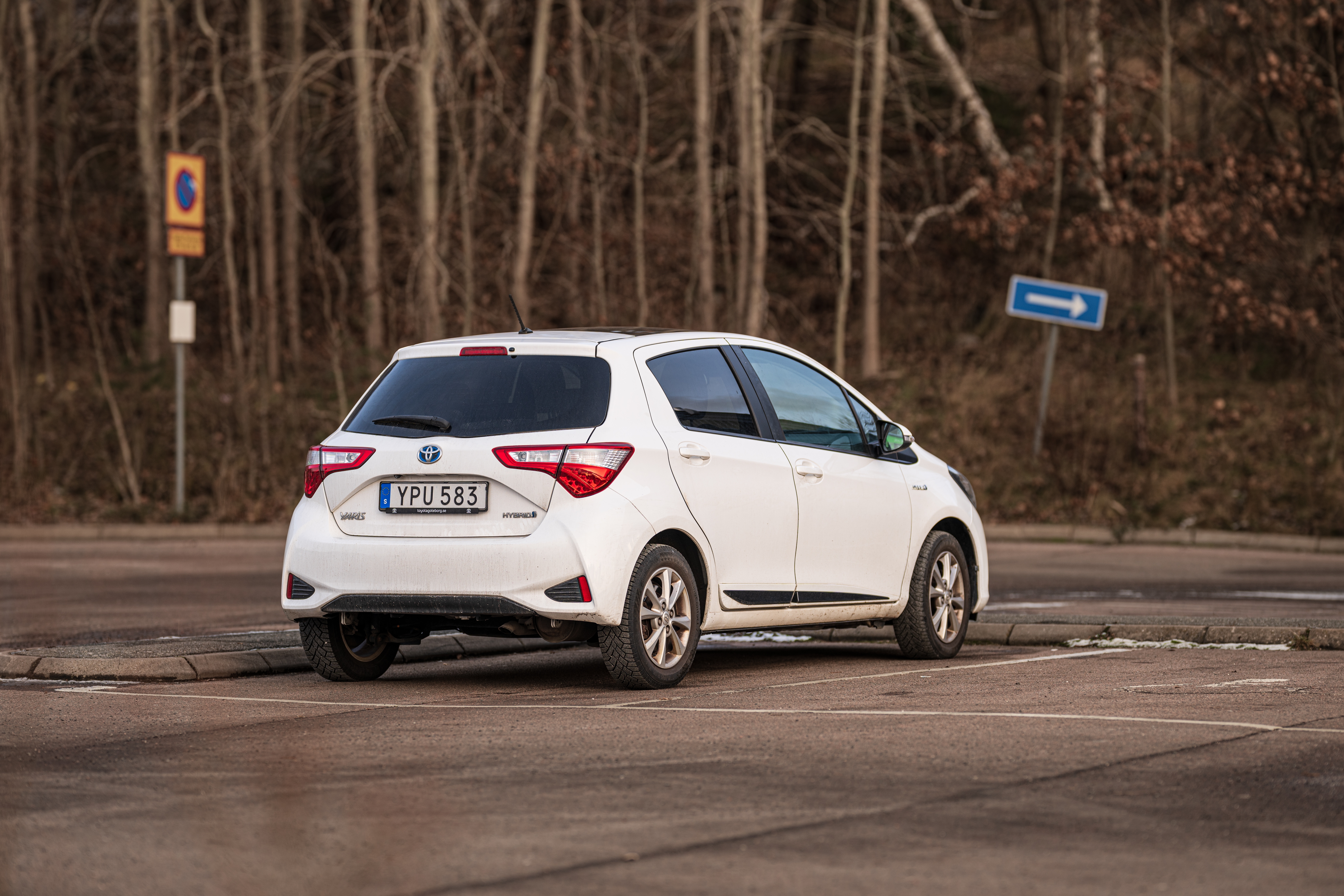 En Toyota Yaris Hybrid står parkerad på en nästan tom parkeringsplats nära en trottoarkant. I bakgrunden finns träd utan löv och trafikskyltar.