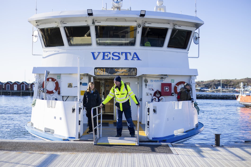 Två personer står vid ingången till en förtöjd färja som heter "VESTA". I bakgrunden syns vatten, båtar och byggnader.