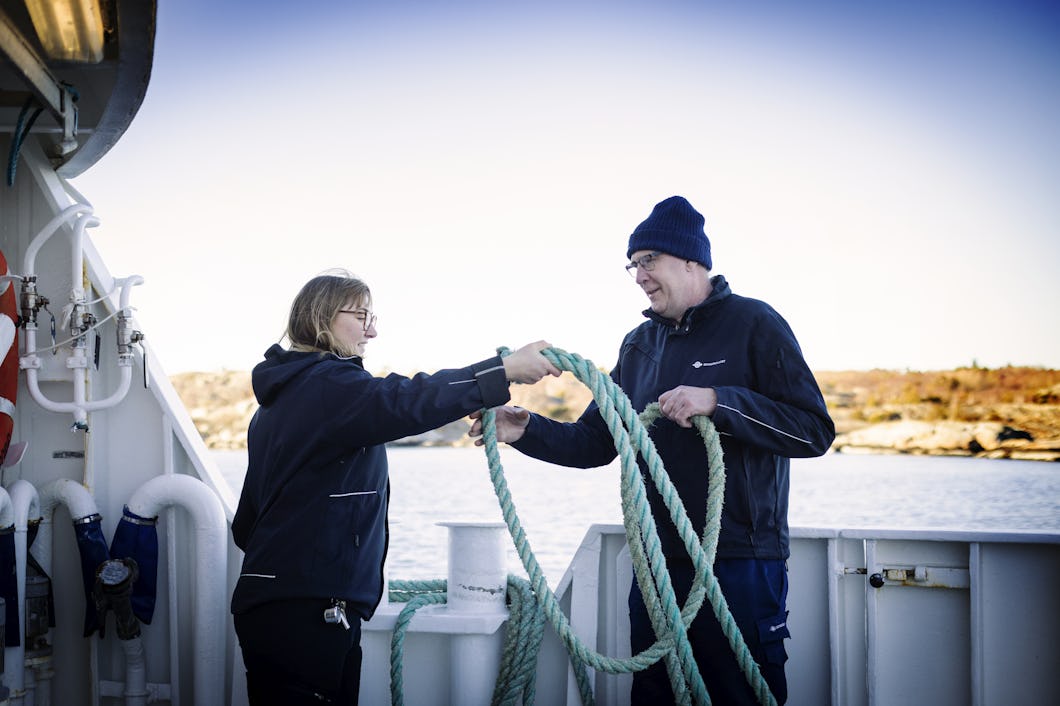 Två personer på en båt samarbetar för att hantera ett stort grönt rep, med strandlinje och vatten i bakgrunden.