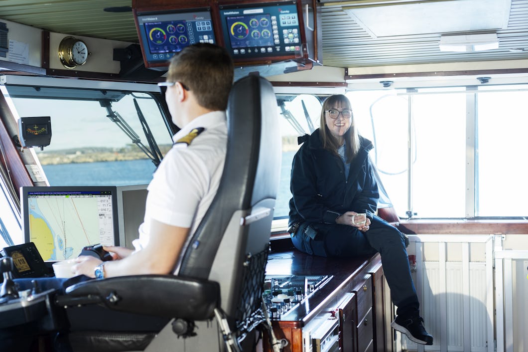En person i uniform styr kontrollerar på ett fartygs brygga medan en annan person sitter på en bänk intill och ler. Navigationsutrustning syns runt dem.