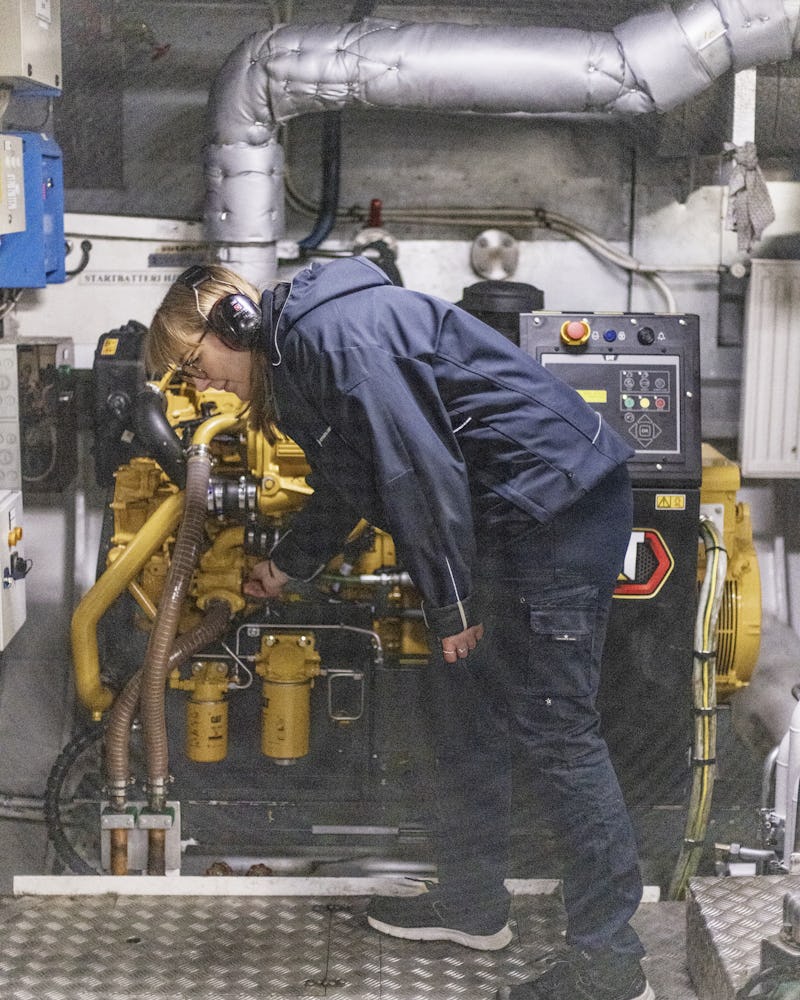 En person med hörlurar och arbetskläder undersöker eller justerar en gul motor i ett industri- eller maskinrum.