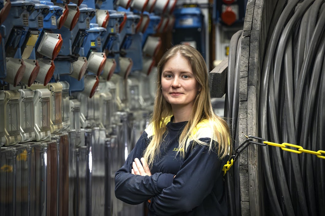 En kvinna med långt blont hår och arbetströja i marinblått står med armarna i kors mellan industriell elutrustning och stora kablar.