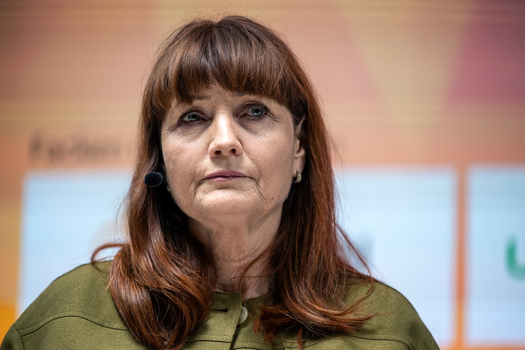 Marie Nilsson med långt brunt hår, iklädd en grön topp och ett headset med mikrofon, står framför en suddig bakgrund i orange och vitt.