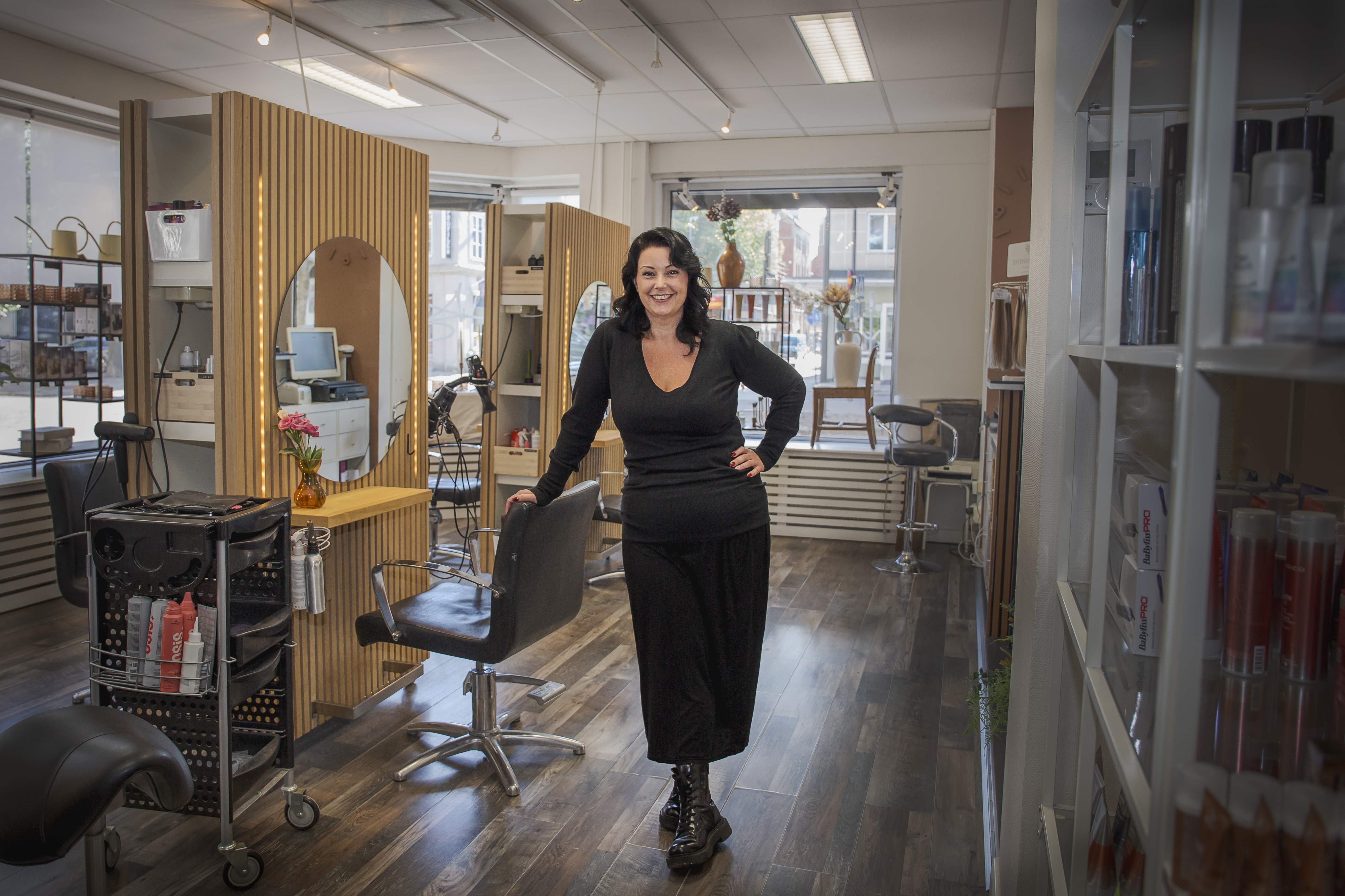 En kvinna står och ler i en frisörsalong med tomma stolar och hårprodukter synliga på hyllor och bänkar.