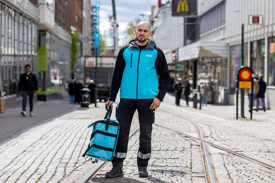 Ammar klädd i en turkos och svart jacka sitter på en betongbänk i ett stadsområde där det finns byggnader och människor i bakgrunden.