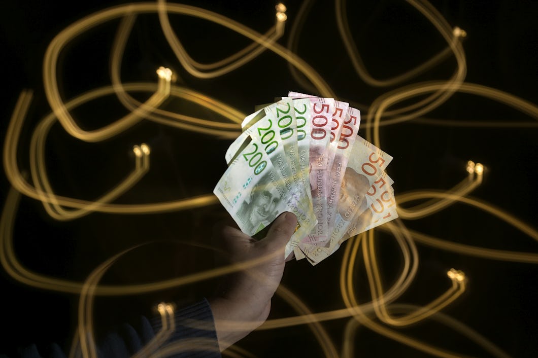En hand håller sedlar av svenska kronor i olika valörer, medan ljusspår syns i bakgrunden mot mörker.