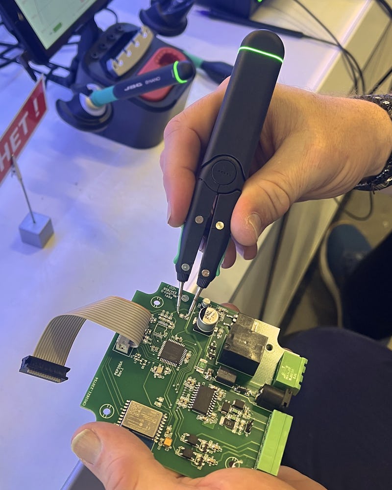 En person använder elektroniska pincetter för att testa komponenter på ett kretskort vid en arbetsstation med verktyg i bakgrunden.