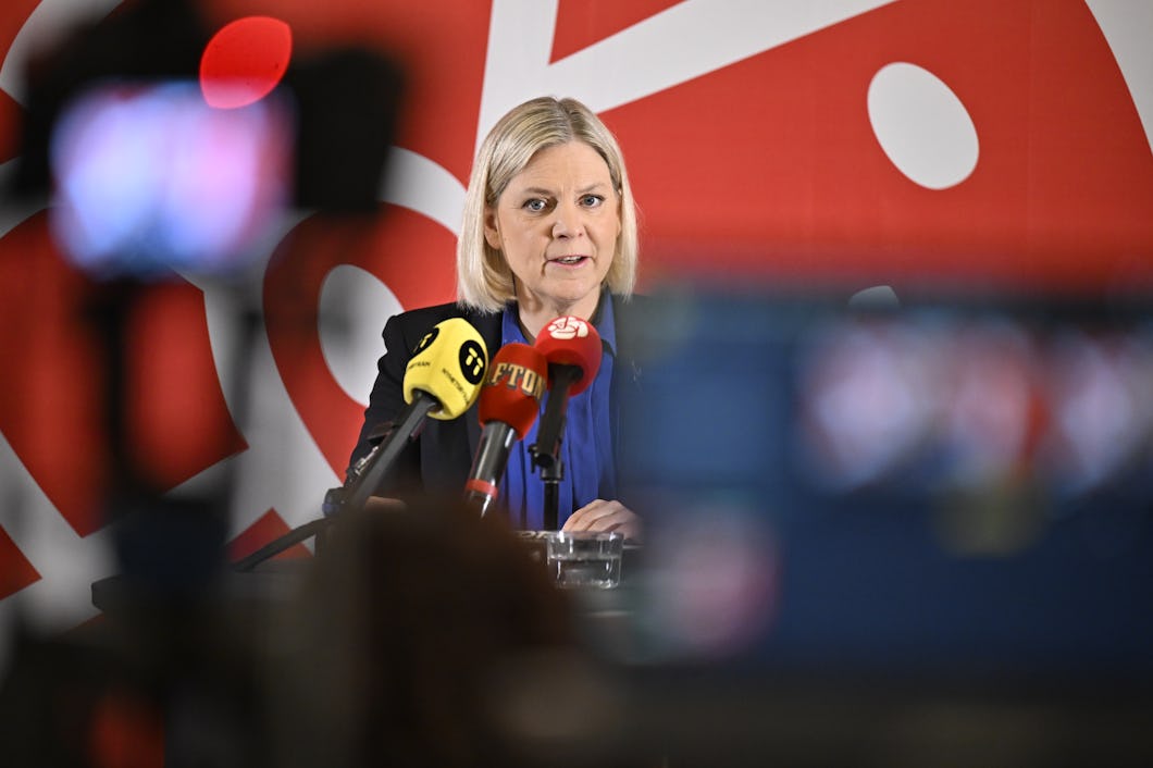 En kvinna talar vid en presskonferens och står bakom flera mikrofoner. Bakom henne finns en rödvit bakgrund med abstrakta mönster.