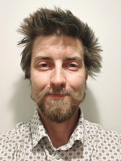 Anton Lorentzon med brunt rufsigt hår, mustasch och skägg står framför en ljus enfärgad vägg och bär en mönstrad skjorta med knappar.