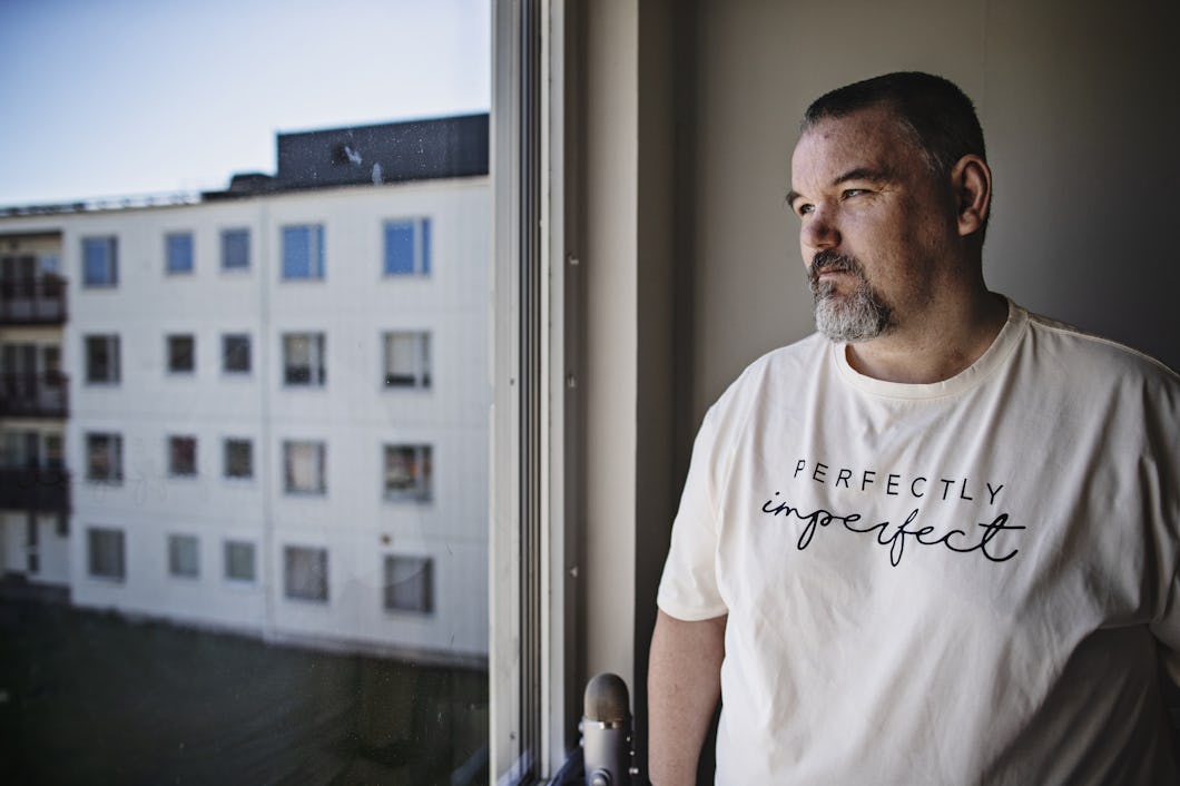 Jimmy Steffenssen med skägg står vid ett fönster och tittar ut. Han har på sig en vit T-shirt med texten ”Perfectly imperfect”. Utanför fönstret syns ett bostadshus.