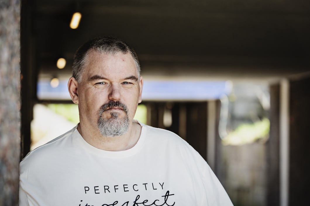 Jimmy Steffenssen med kort grått hår och getskägg står utomhus i skuggan och har på sig en vit t-shirt med tryckt text.