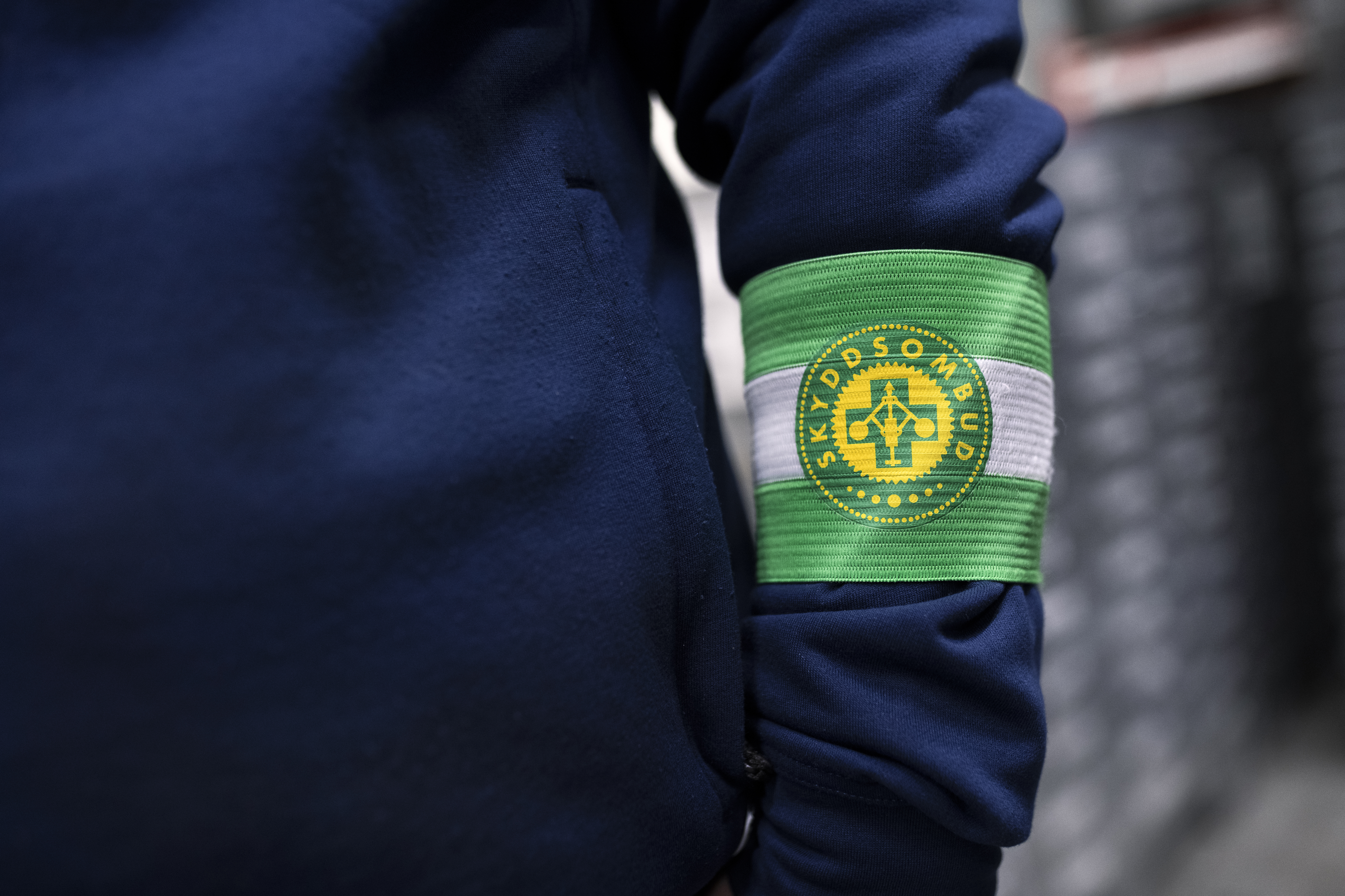 En person bär en jacka och har ett grönvitt armband med texten "SKYDDSOMBUD" och en logotyp.
