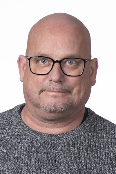 Håkan Hallengren med rakat huvud, glasögon och kort skägg har på sig en stickad tröja och vänder sig mot kameran framför en vit bakgrund.