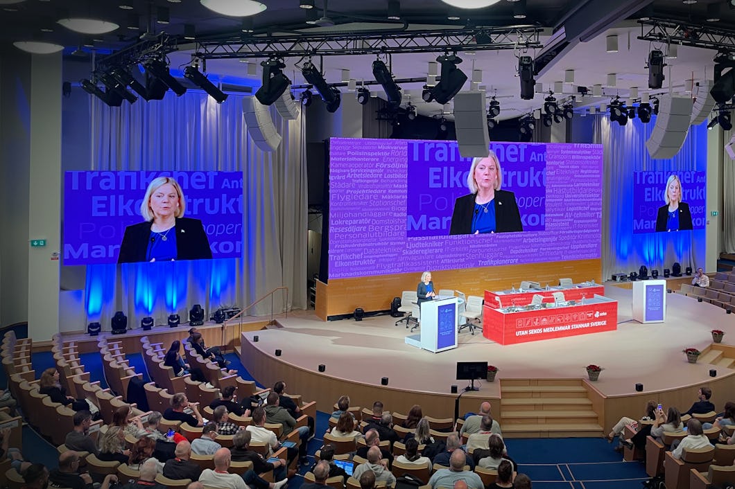 En kvinna talar vid ett podium i en aula inför en publik. Hennes bild visa på en skärm bakom henne med blått och lila ljus.