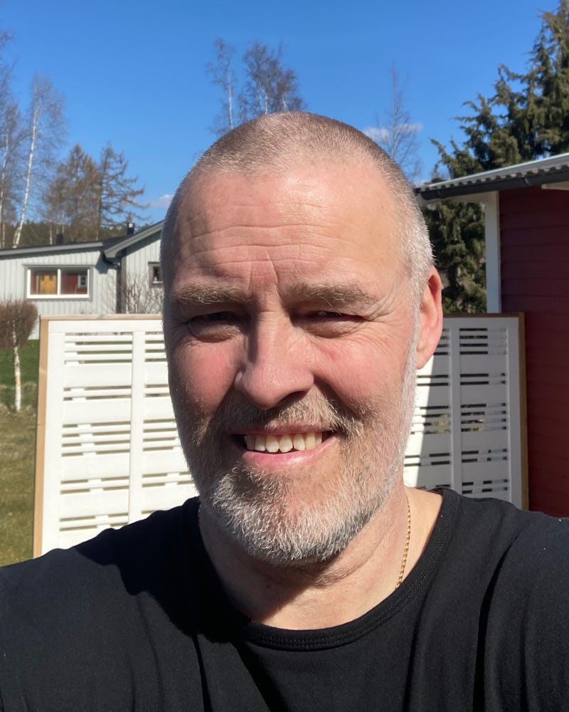 En medelålders man med snaggat hår och skägg ler utomhus. I bakgrunden syns hus, träd och ett vitt staket.