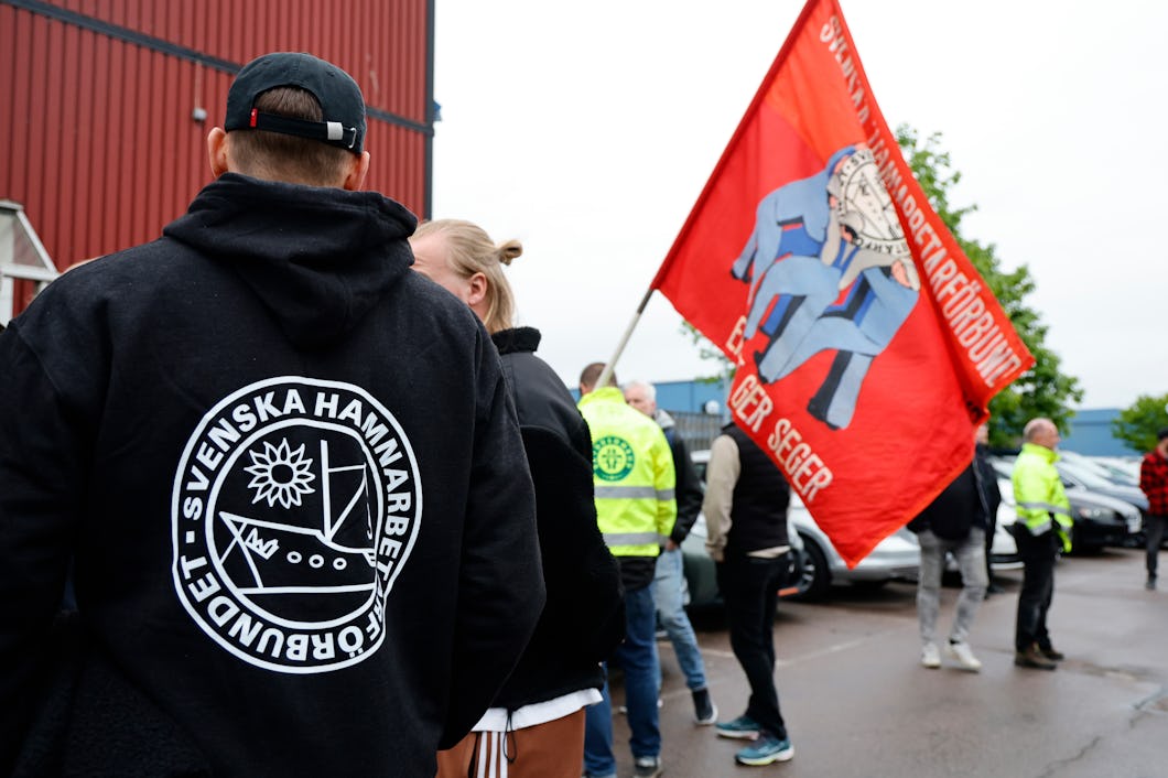 Personer står utomhus, några har jackor med texten "Svenska Hamnarbetarförbundet", medan andra håller i en röd flagga med text och en bild. De befinner sig nära parkerade bilar och byggnader.