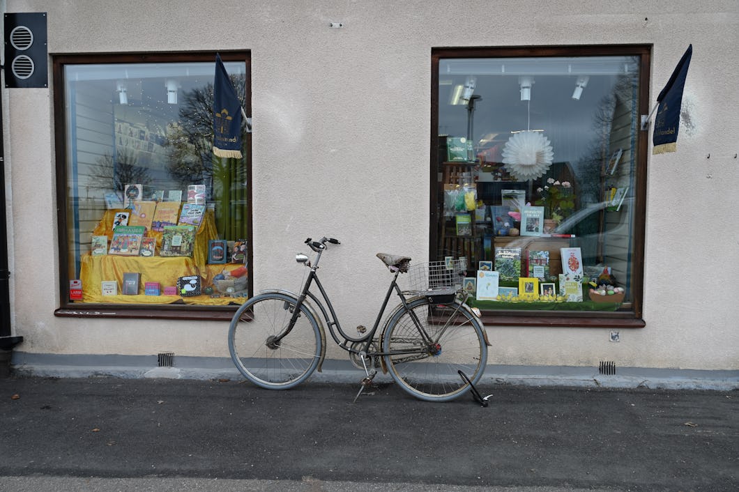 En cykel står parkerad på trottoaren framför en butik med två stora skyltfönster där böcker och föremål visa.