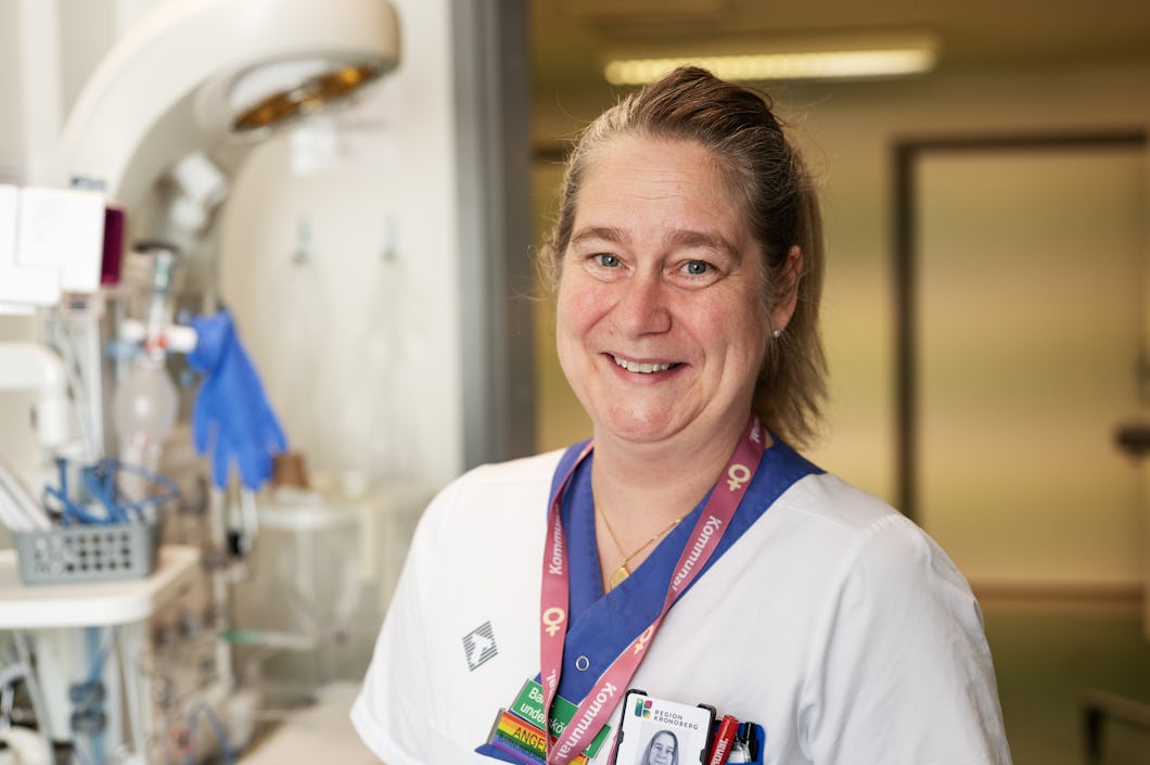 Angelica Robertsson med arbetskläder och namnskylt står i en medicinsk miljö där utrustning syns i bakgrunden.