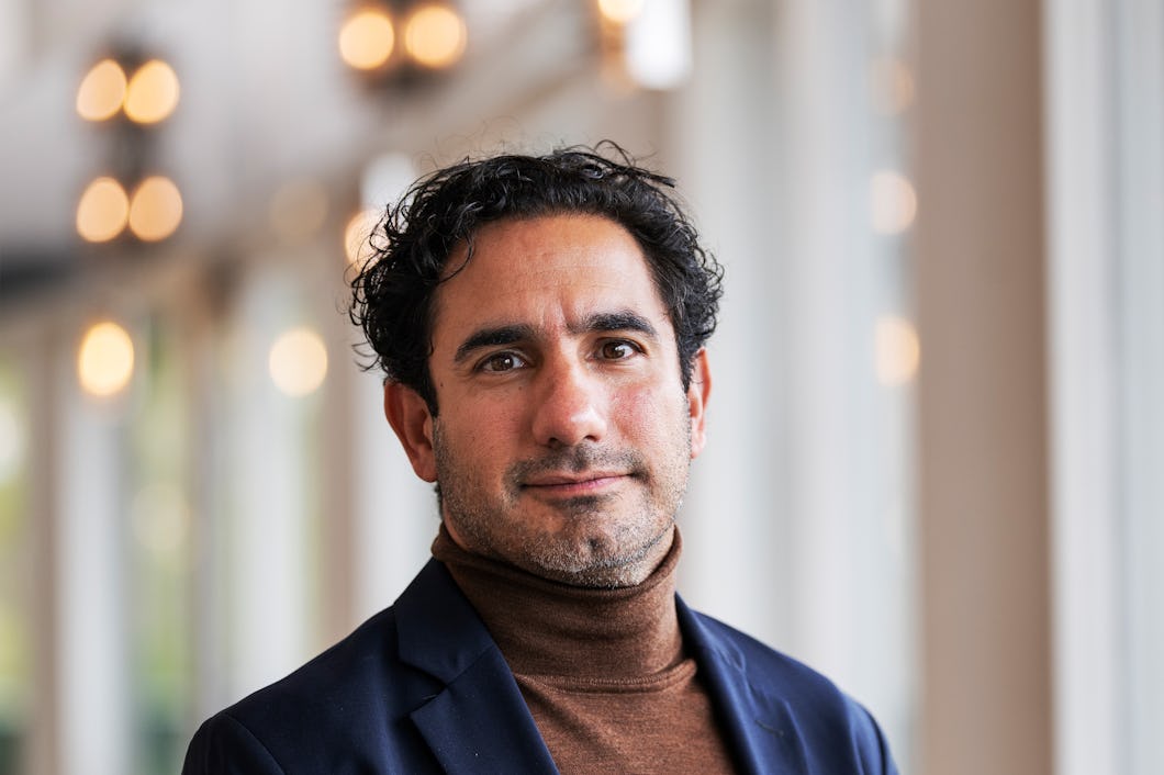 Ardalan Shekarab med lockigt mörkt hår, klädd i polotröja och kavaj, står inomhus nära stora fönster med suddiga ljus i bakgrunden.