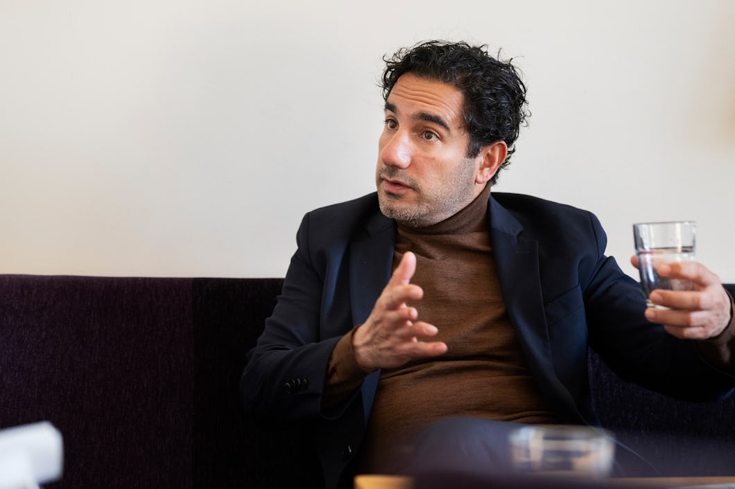Ardalan Shekarabi med lockigt hår sitter på en soffa. Han gestikulerar med ena handen och håller ett glas vatten i den andra. Han har på sig kavaj och polotröja.