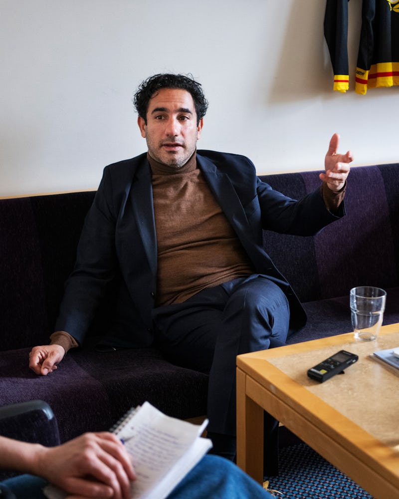 En man i kostym sitter på en soffa och gör en handrörelse. En annan person i närheten håller i ett anteckningsblock. På bordet finns ett glas vatten och en inspelningsapparat.