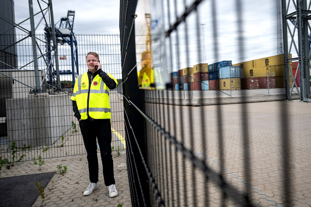 En man med en gul säkerhetsväst står vid ett metallstaket på en lastplats och talar i telefon. I bakgrunden syns containrar och lyftkranar.