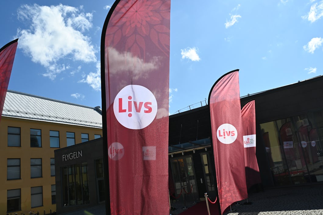 Röda flaggor med Livs logotyp står utanför en byggnad med gul fasad och ordet "Flygeln" på väggen under en himmel med moln.
