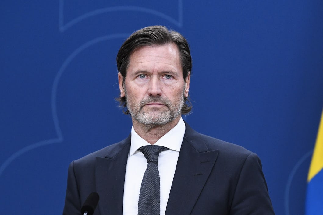 En man i kostym och slips står vid en talarstol med neutralt ansiktsuttryck.
