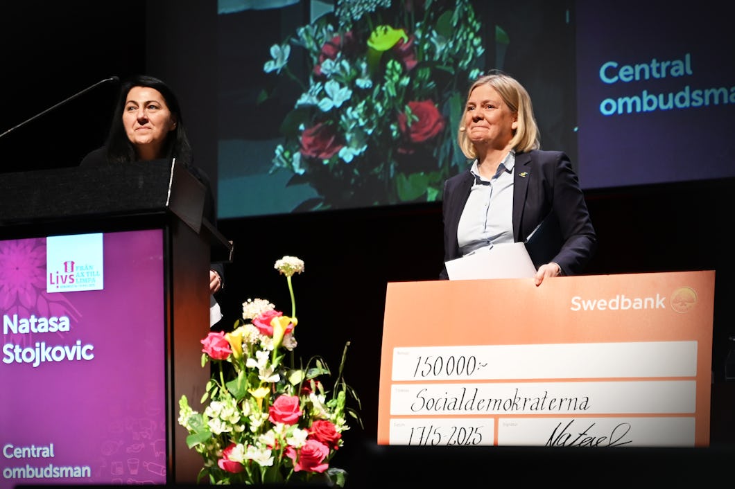 Två kvinnor står på scen. Den ena talar vid ett podium och den andra håller i en stor check. I bakgrunden finns blommor.