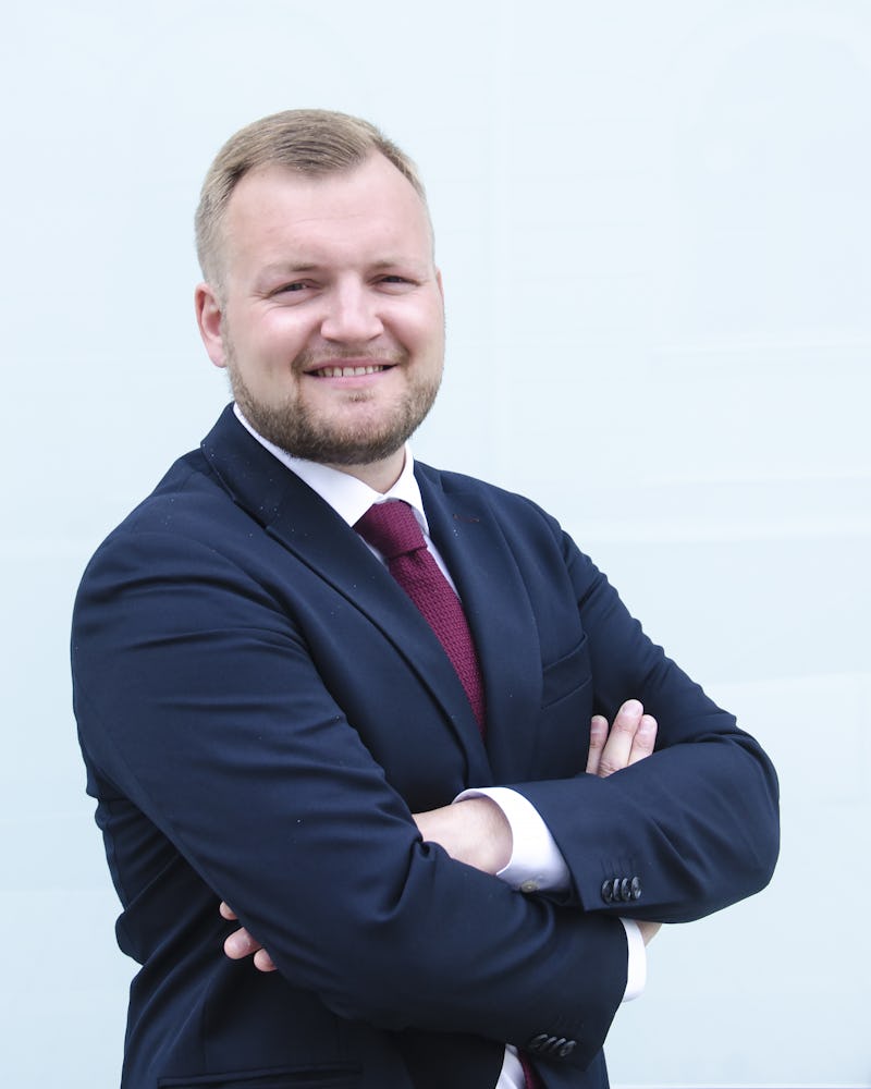 Erik Sinander i mörkblå kostym och röd slips, står med armarna i kors och ler mot kameran framför en ljus bakgrund.