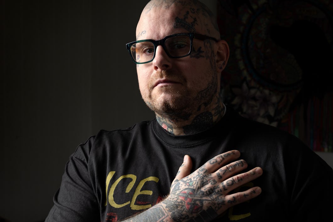 David Gustavsson med mössa och glasögon står utomhus vid solnedgången, med hus i bakgrunden. Han har på sig en jacka med ett märke och håller en hand på bröstet. På handen saknas toppen på ringfingret.