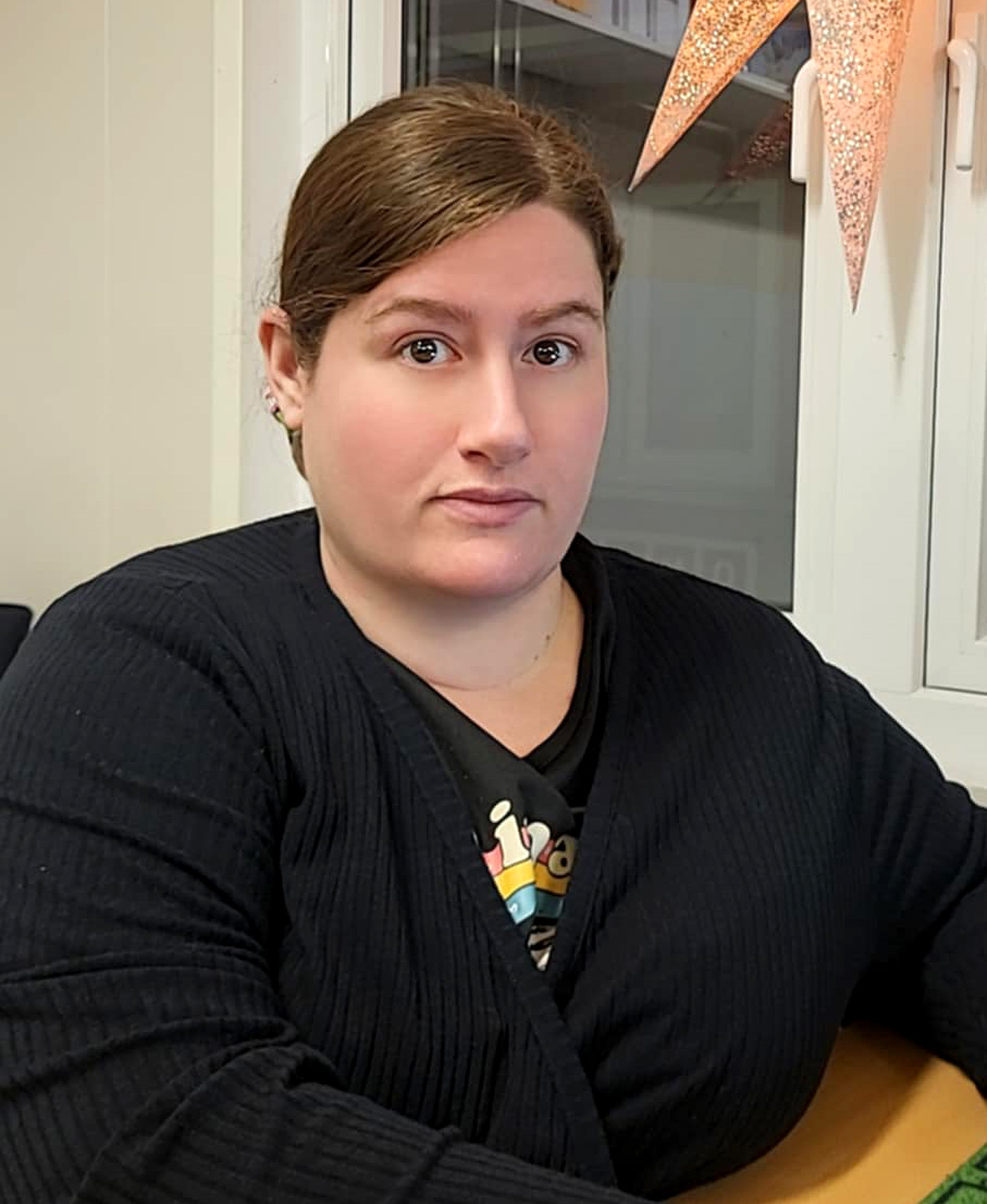 Victoria Hart med brunt hår sitter inomhus vid ett fönster. Hon har på sig en svart kofta och en mönstrad skjorta och tittar rakt mot kameran.