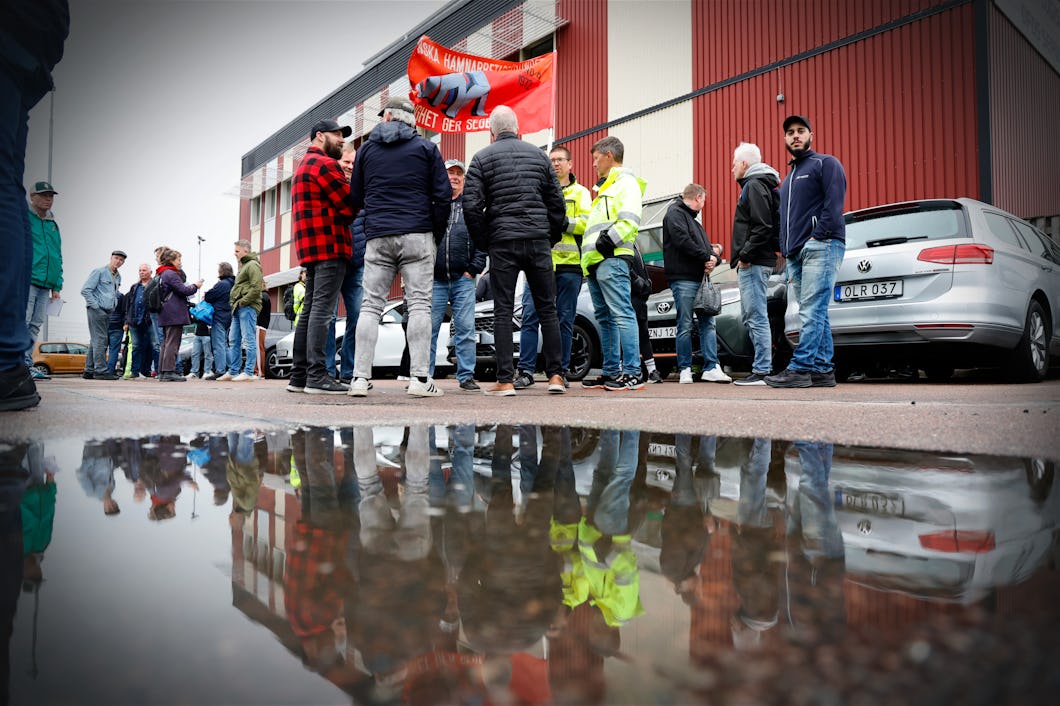 En grupp personer står och pratar utanför en byggnad nära parkerade bilar. Några av dem håller i en röd banderoll. Deras spegelbild syns i en stor vattenpöl.