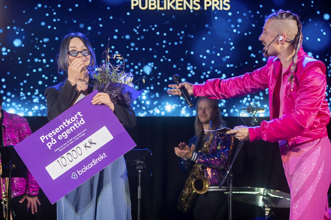 En kvinna håller blommor och en stor check på 10 000 kronor när hon tar emot ett pris på scenen. En person i rosa kläder delar ut priset och applåderar.