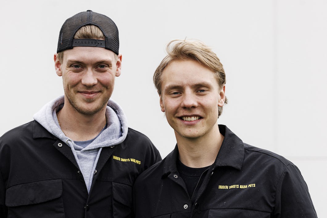 Två män i svarta jackor står bredvid varandra framför en vit bakgrund. En av dem har en keps bak och fram.
