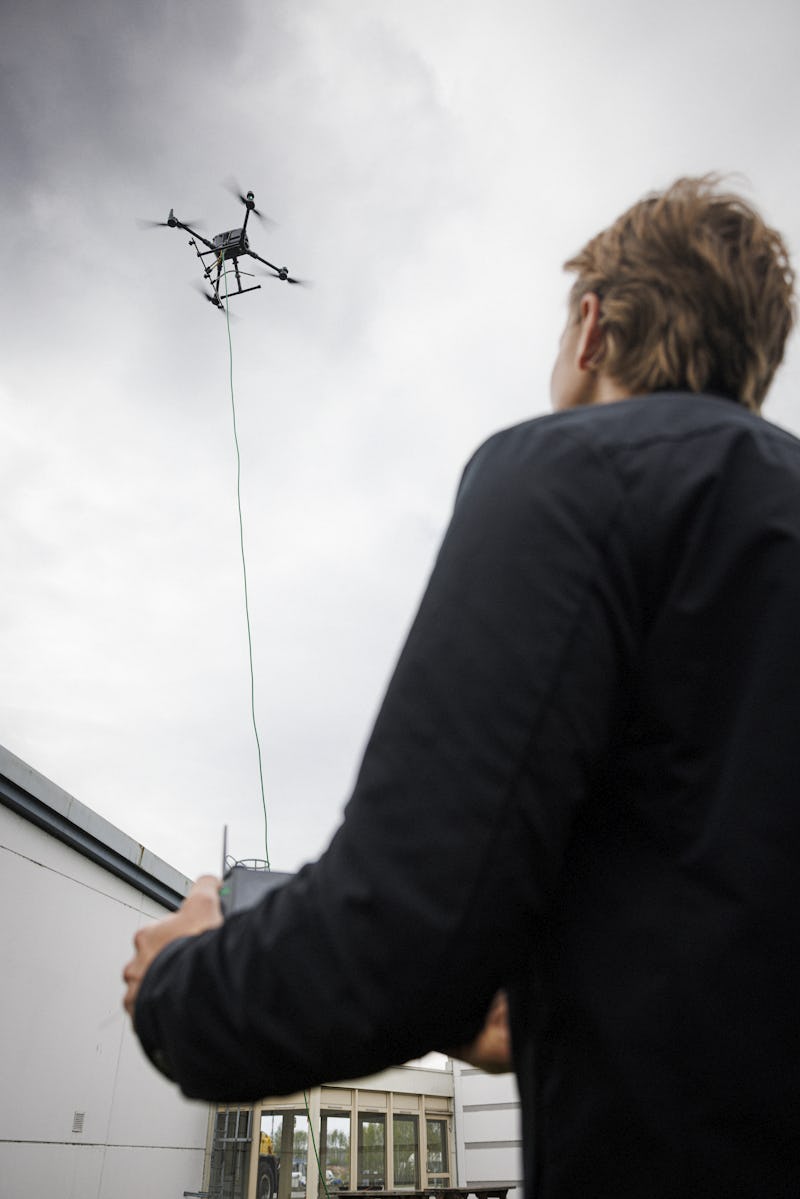 En person styr en drönare som är kopplad med en kabel och flyger nära en byggnad under molnig himmel.
