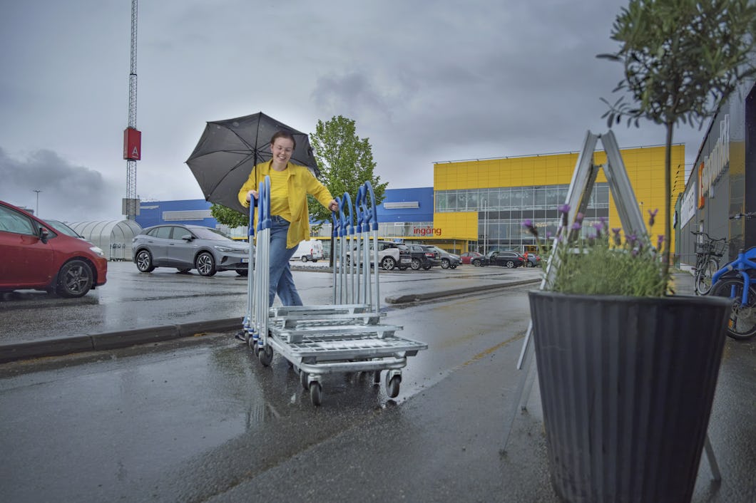 En person bär en gul jacka och håller i ett ut-och-inväntat paraply samtidigt som han skjuter kundvagnar utanför en stor butik med gula och blå färger under regnet.