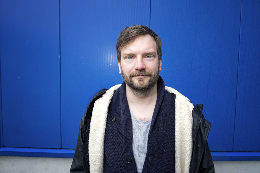 Karl Segerquistmed skägg och mustasch står framför en blå vägg. Han har på sig en grå skjorta, en mörk scarf och en svart jacka med ljus insida.