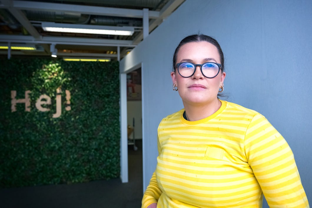 Kajsa Lindgren i randig gul skjorta och runda glasögon står inomhus nära en vägg täckt av växter med ordet "Hej!".