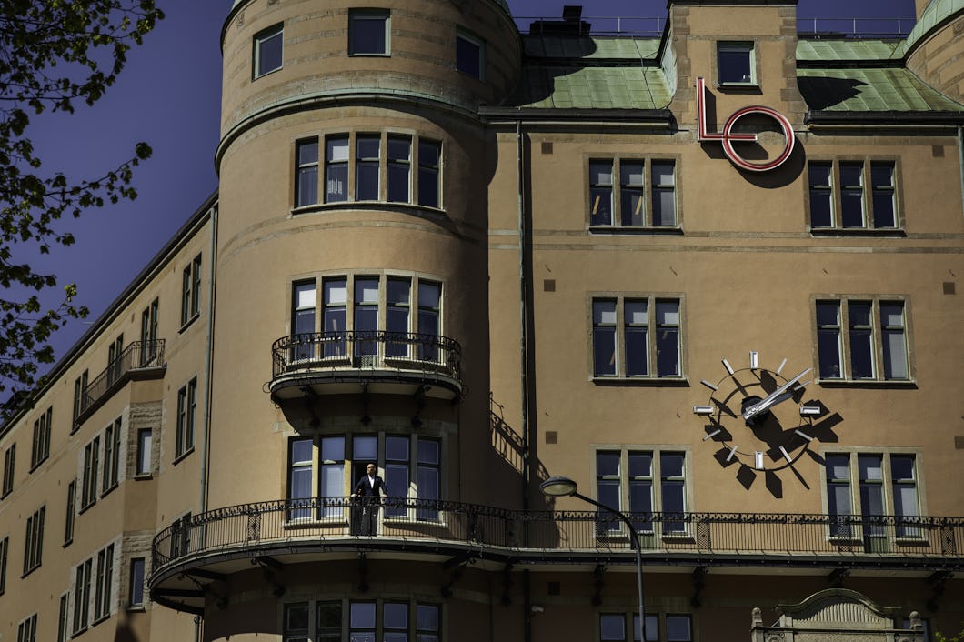En person står på en balkong som tillhör en byggnad med många fönster, en klocka och röda bokstäverna "LO" på taket.