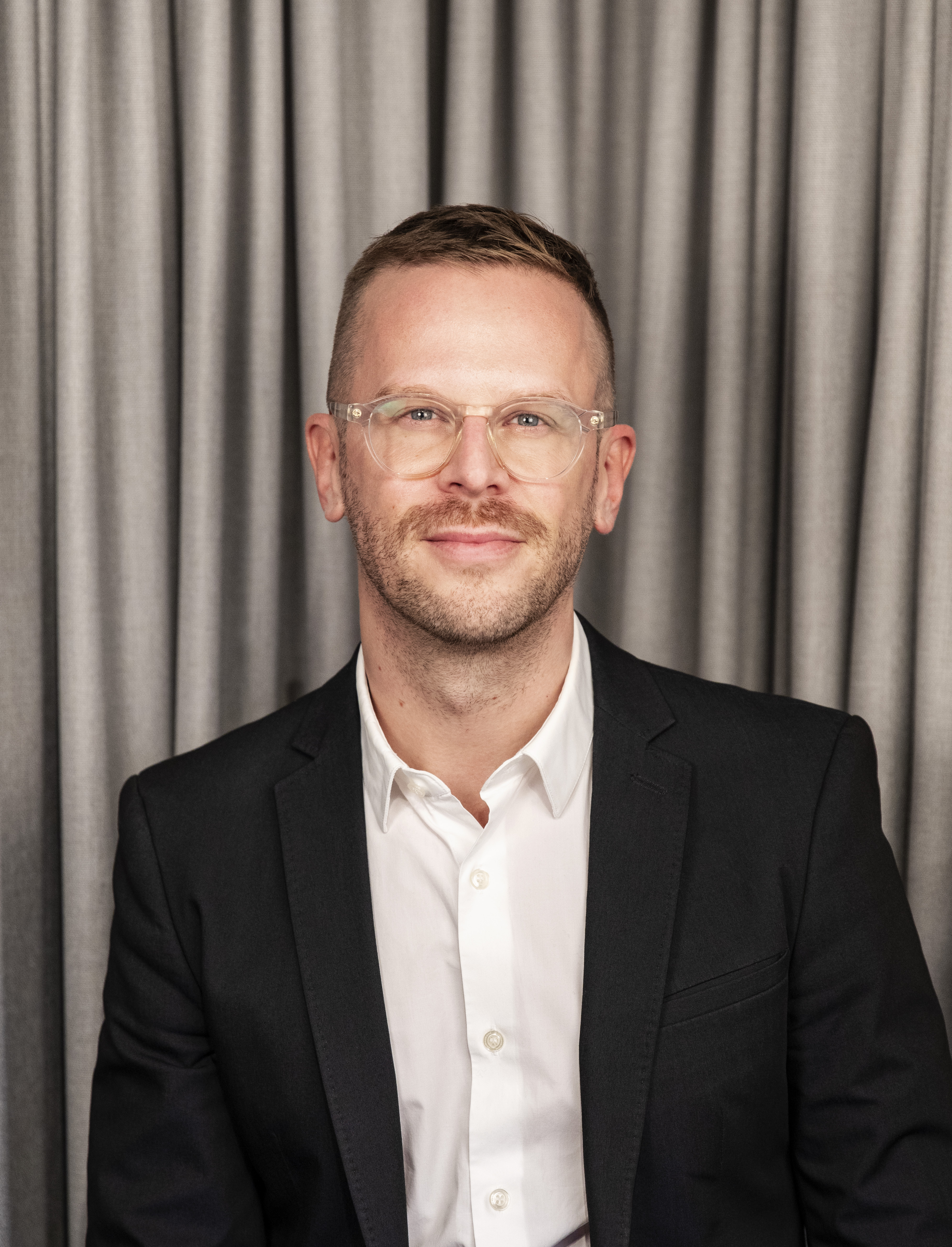 Daniel Pus med kort hår, glasögon och skägg sitter i svart kostym och vit skjorta framför grå gardiner.