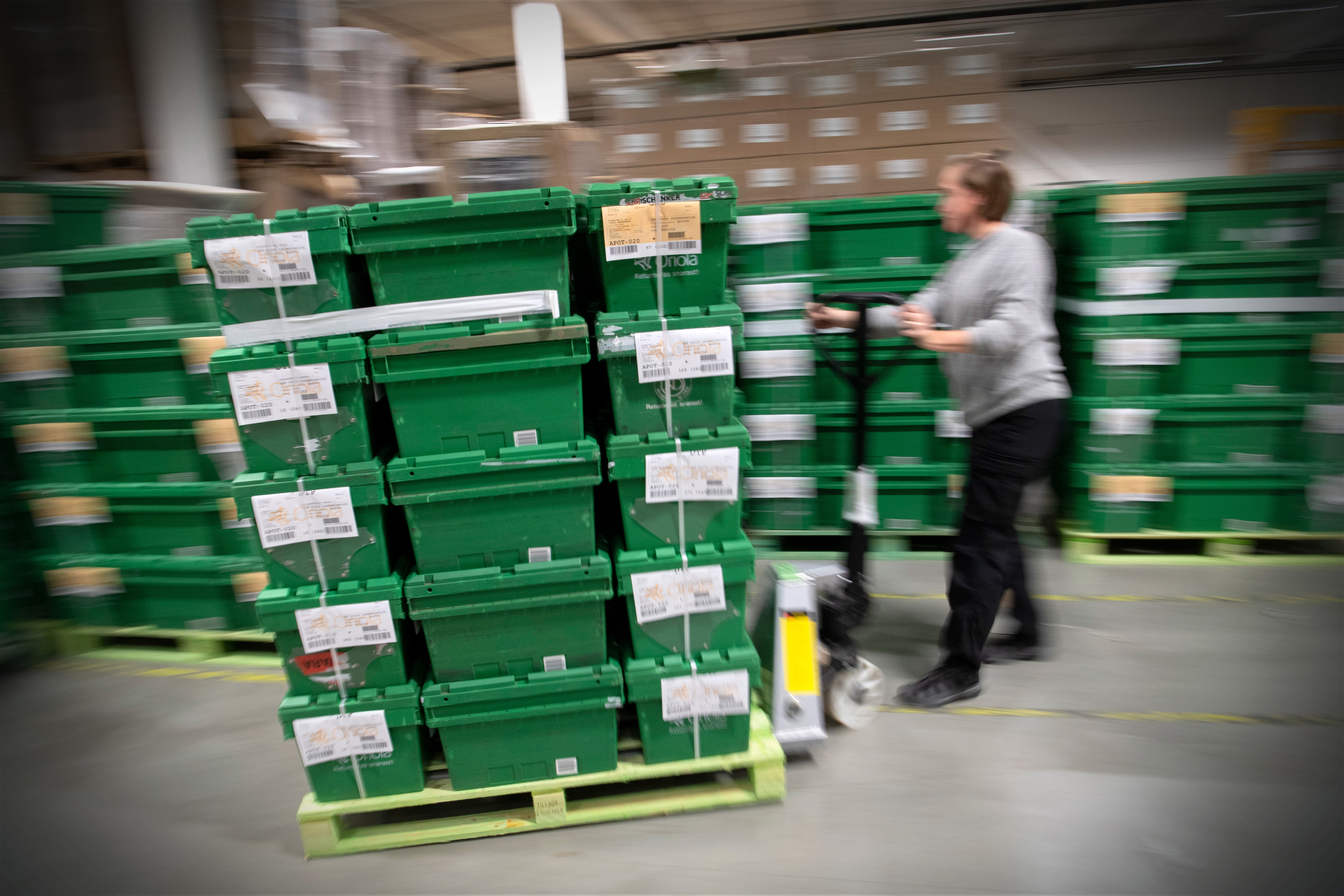 En person skjuter en vagn med staplade plastlådor i ett lager. I bakgrunden syns hyllor och kartonger.