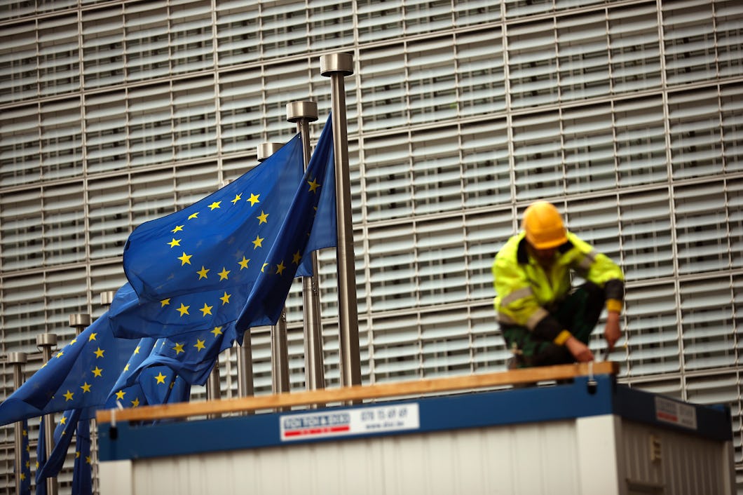 En arbetare med hjälm arbetar nära EU-flaggor framför en byggnad med metallfasad.