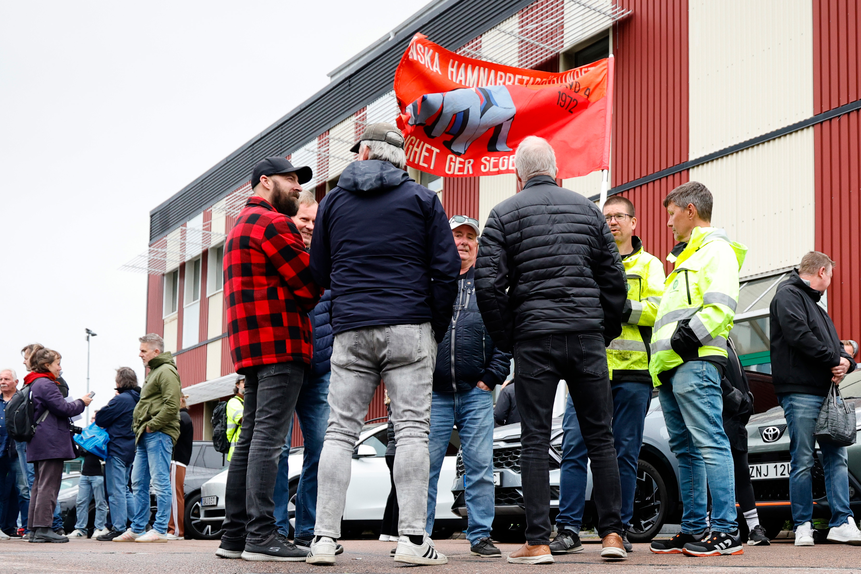En grupp personer står och pratar utanför en byggnad. Två män håller i ett rött plakat med en björn och svensk text.