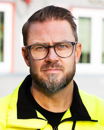 Kim Söderström glasögon och skägg bär en gul varseljacka och står utomhus utomhus en byggnad med fönster.