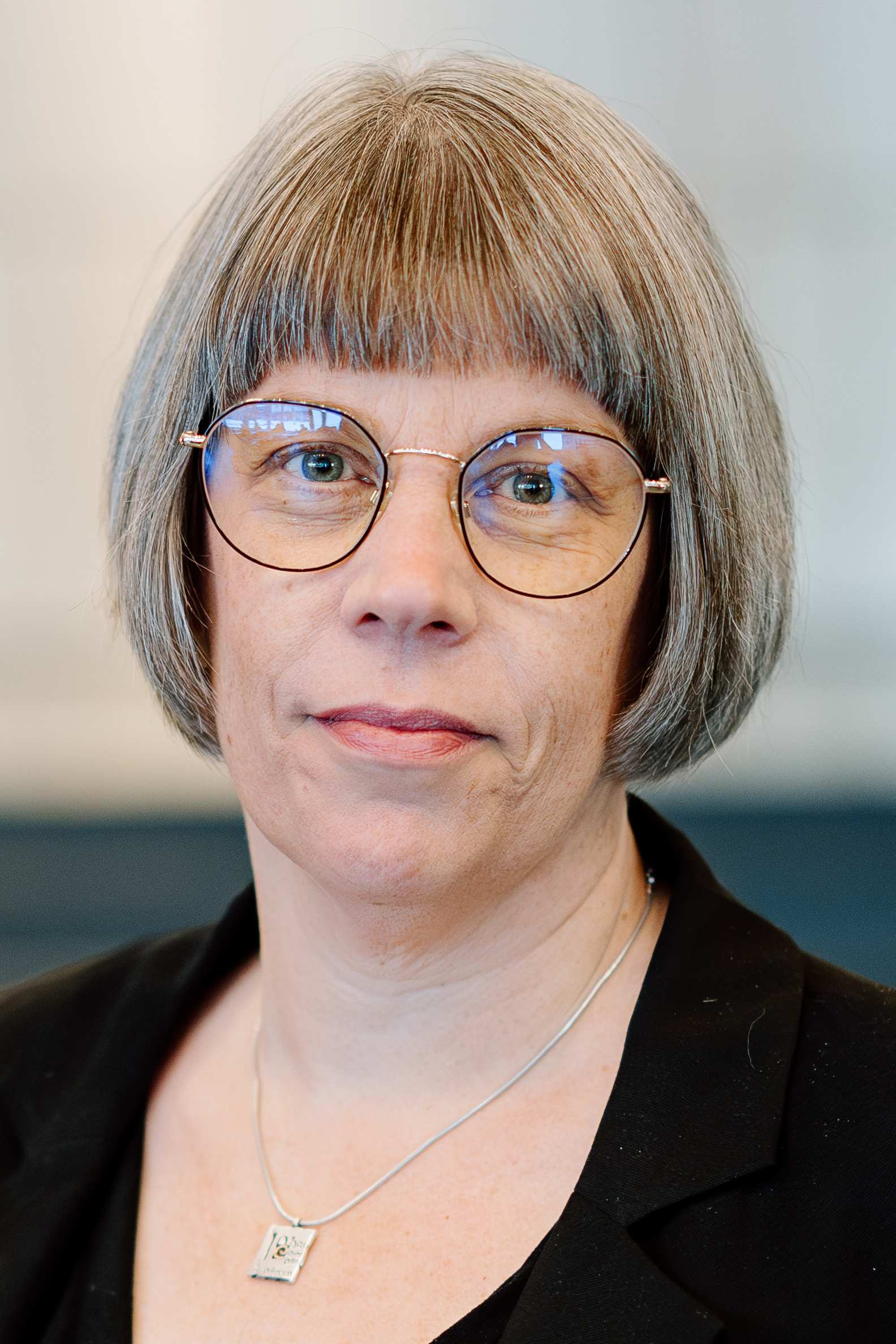 En kvinna med rakt grått hår, glasögon och svart kavaj ser in i kameran med neutral min. Hon har ett halsband med fyrkantigt hänge.