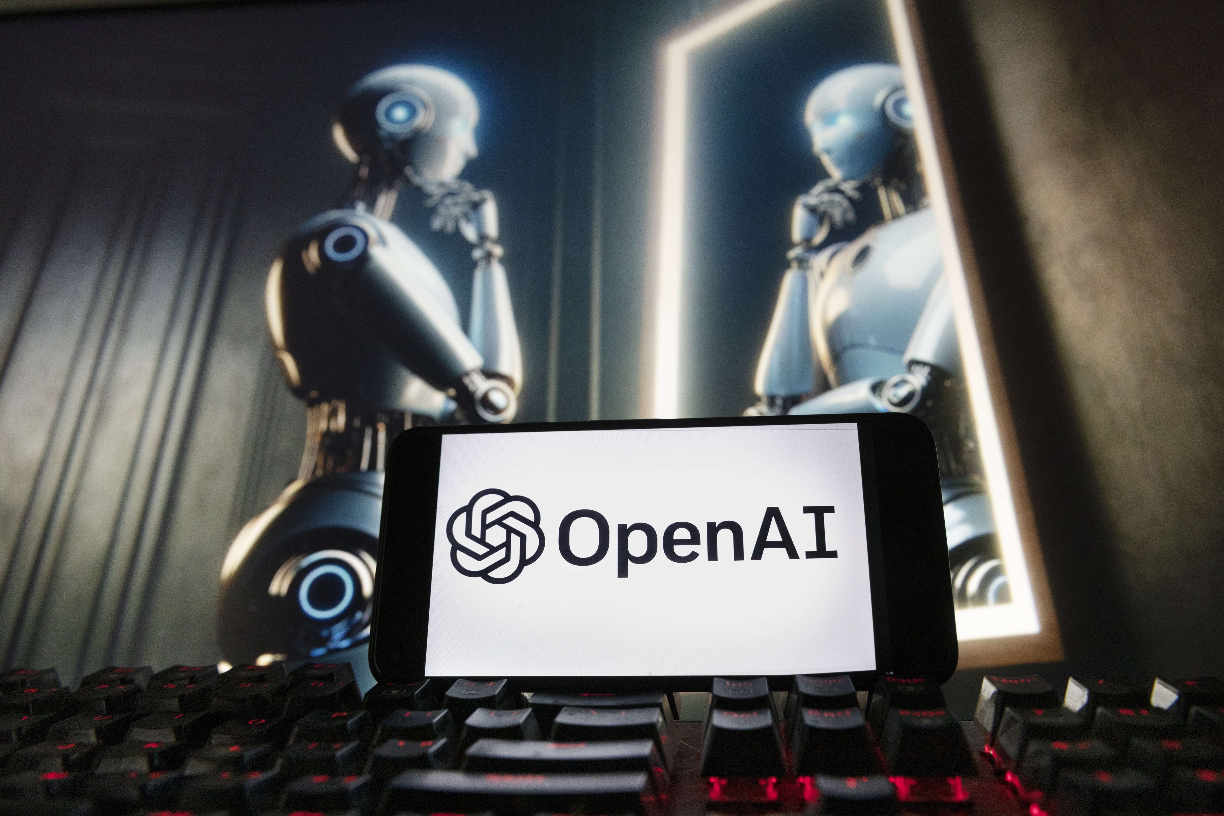 En smartphone med OpenAI:s logotyp ligger på ett tangentbord. I bakgrunden visa en skärm där en människoliknande robot ser sig själv i en spegel.